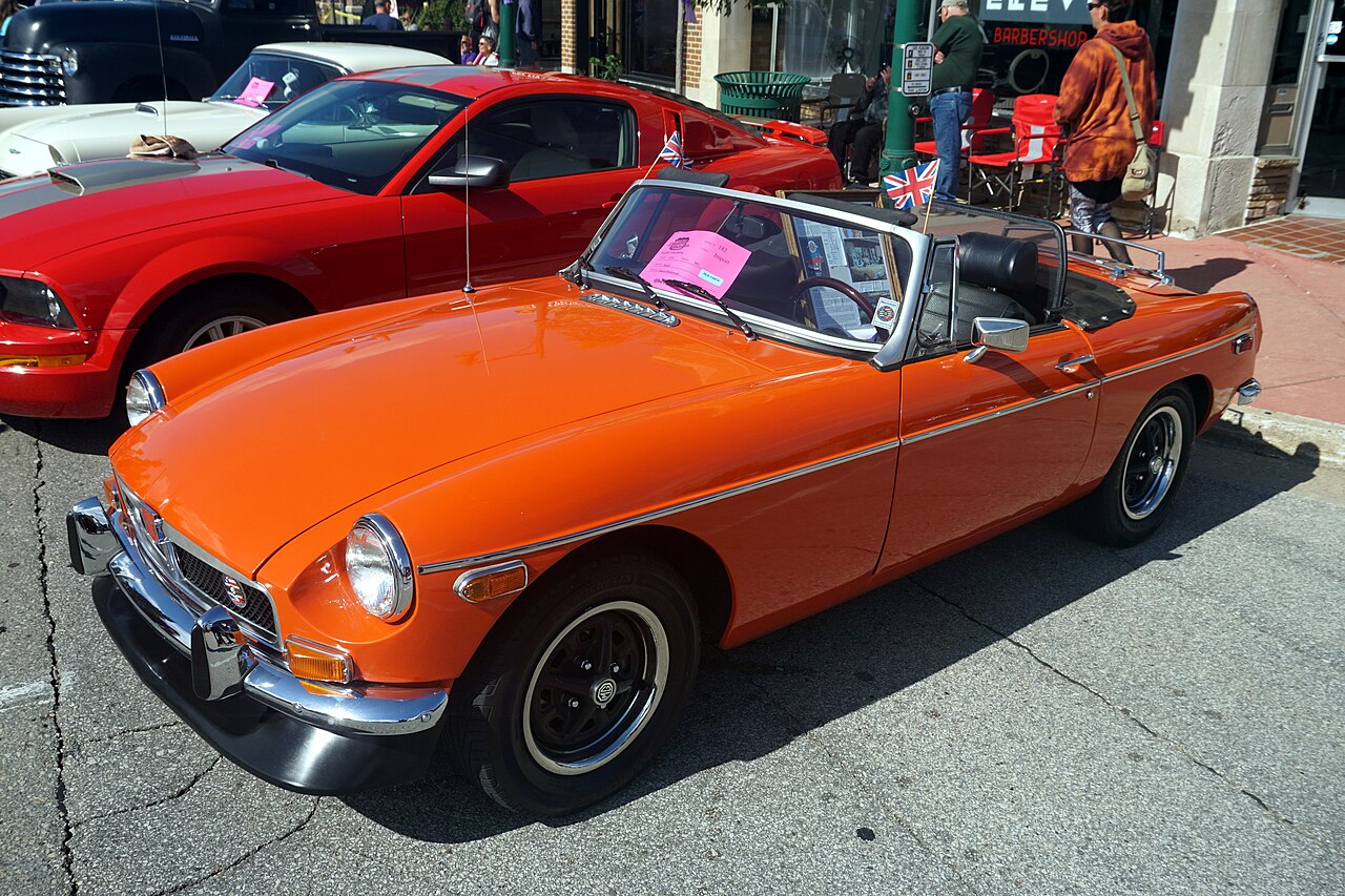 1974 MG MGB roadster in orange with its roof down and two Union Jack flags, front left three-quarter view on a street at a car show.