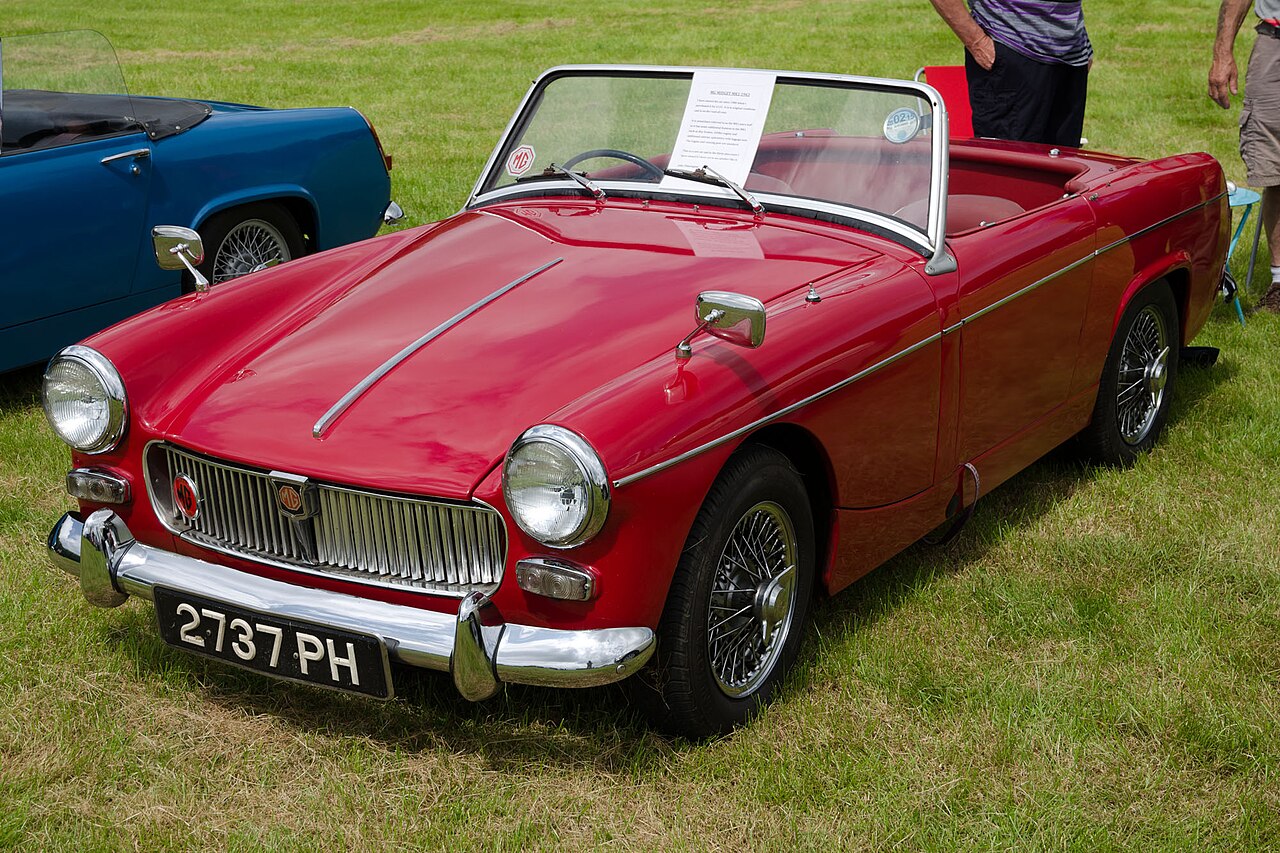Red 1963 MG Midget Mk I with wire wheels and its roof down, front left three-quarter view on grass at a car show.
