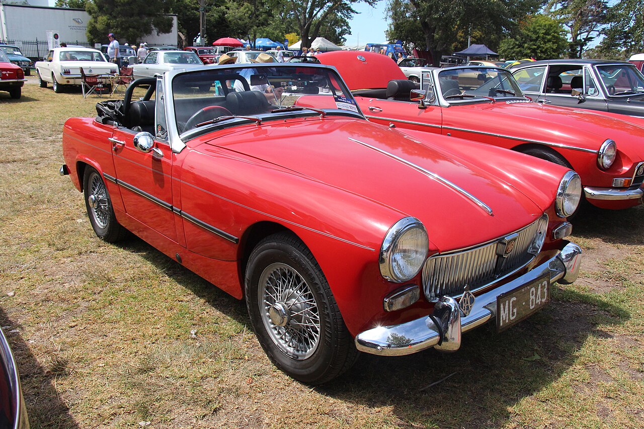 Red 1965 MG Midget Mk II roadster with wire wheels and its roof down, front three-quarter view on grass at a car show.