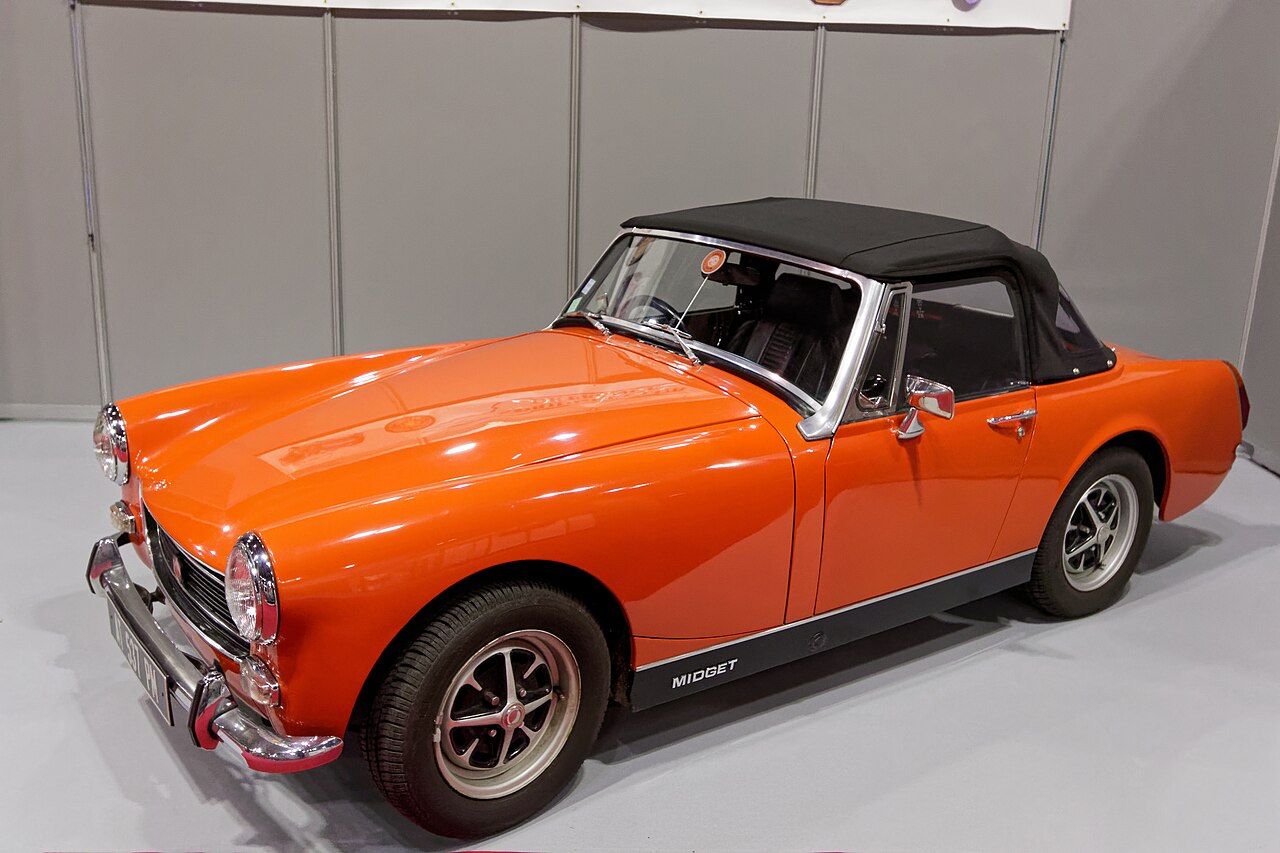 Orange MG Midget roadster with its black soft top up, front left three-quarter view at an indoor classic car show.