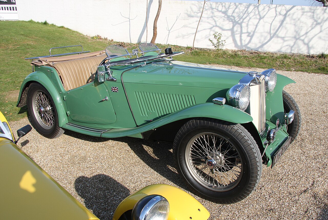 Pale green early MG T-type roadster with wire wheels and its roof down, front right three-quarter view on gravel.