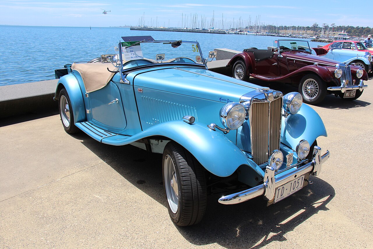 1951 MG TD roadster in pale blue with its roof down, front right three-quarter view on a waterfront promenade.