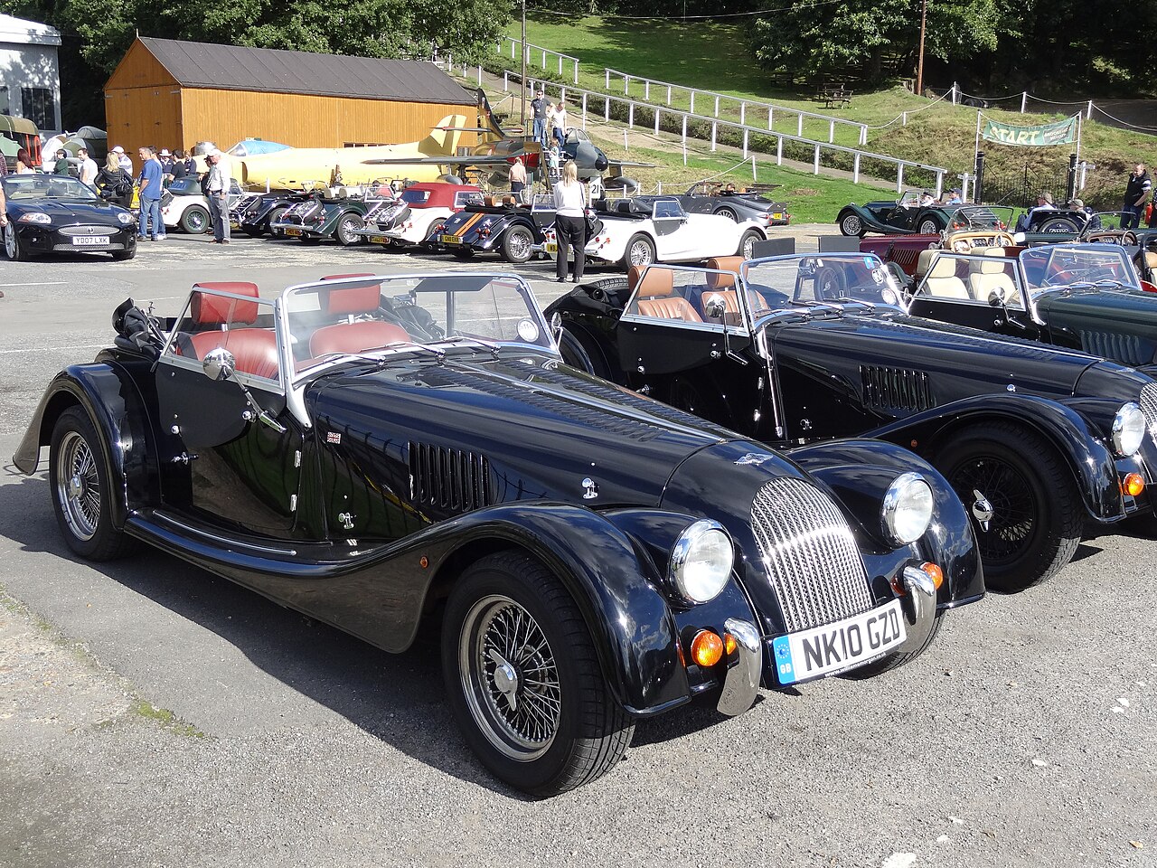 Black Morgan roadster with wire wheels and red seats beside a second black Morgan at a 2013 Brooklands gathering, front three-quarter view.