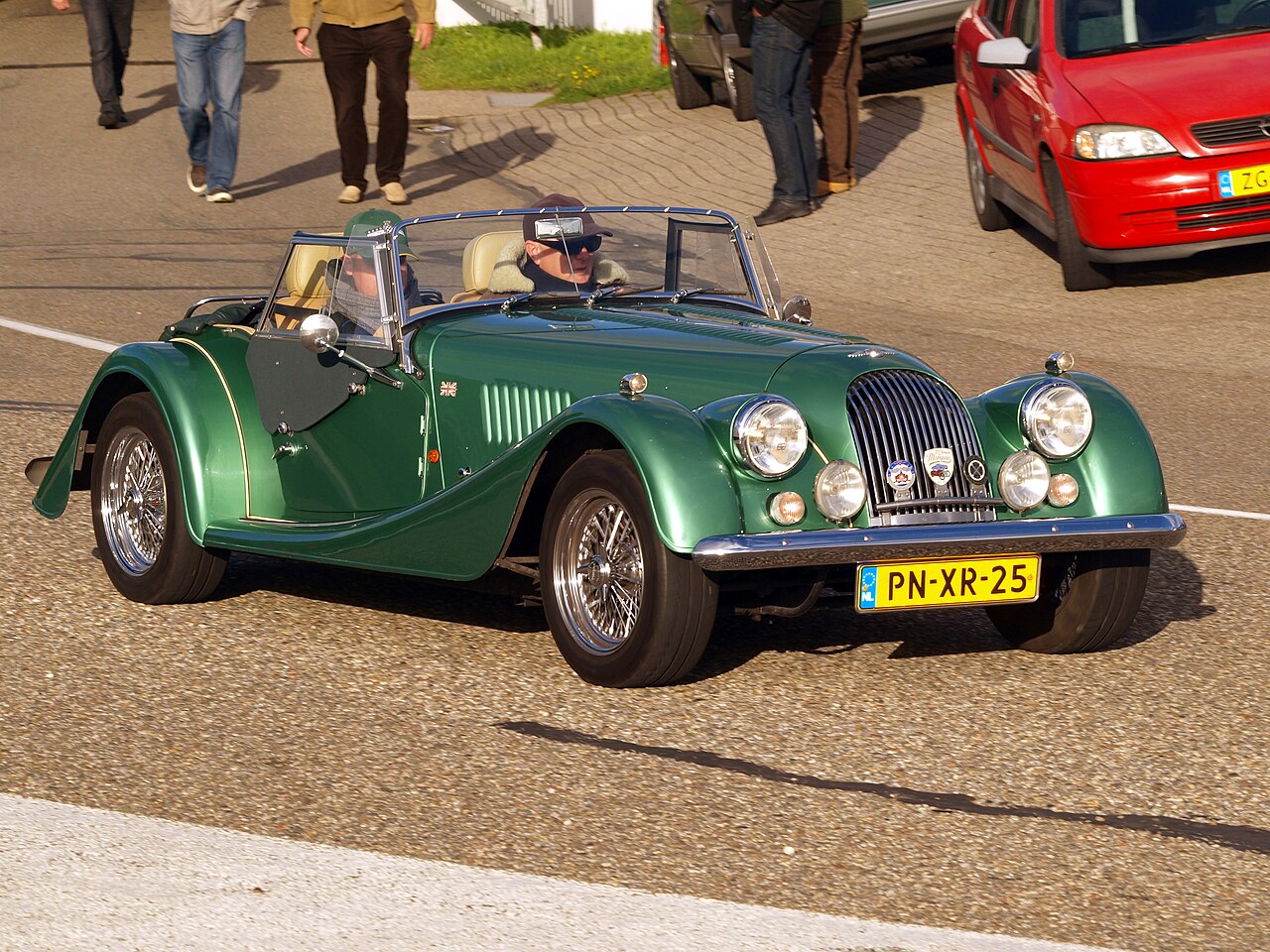 1995 Morgan Plus 4 in green with wire wheels and its hood down, driving past with two occupants, front three-quarter view.