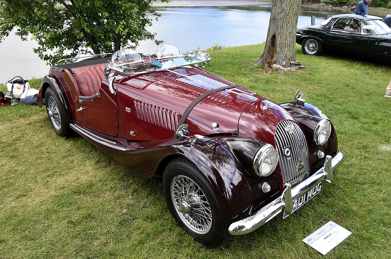 1960 Morgan Plus 4 in burgundy with wire wheels and its roof down, front right three-quarter view on grass beside the water.