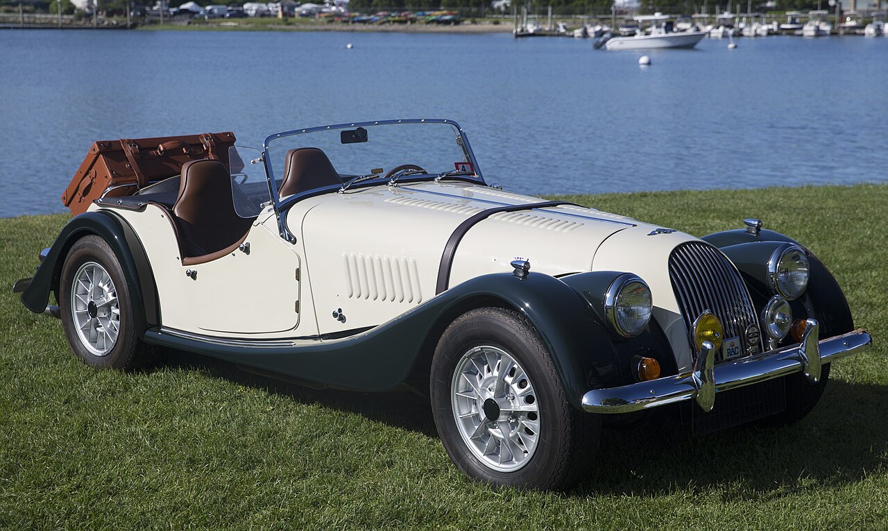 1971 Morgan Plus 8 in ivory with dark green wings and its roof down, front right three-quarter view on grass beside a harbour.