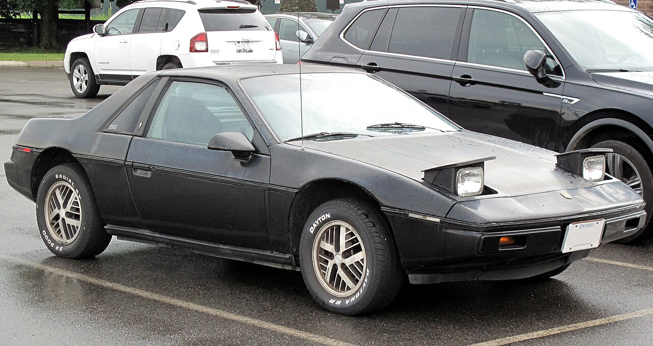 Black 1984 Pontiac Fiero SE with its pop-up headlamps raised, front three-quarter view in a wet car park.