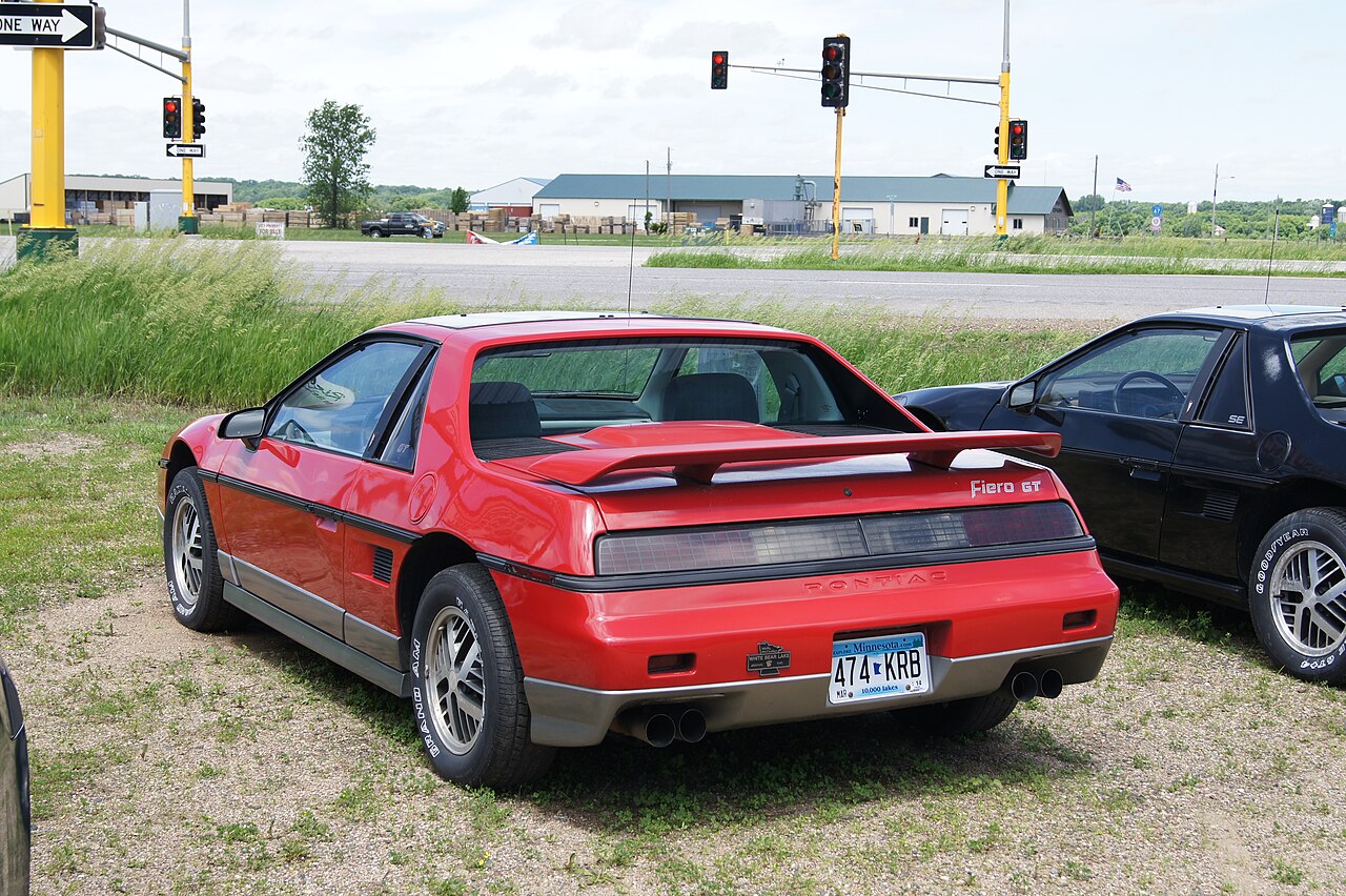 Red 1986 Pontiac Fiero GT with a rear spoiler, rear three-quarter view on grass and gravel beside a black Fiero.