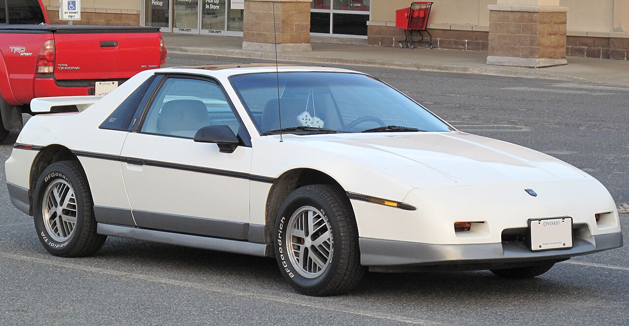 White 1985 Pontiac Fiero GT with fuzzy dice hanging inside, front right three-quarter view parked in front of a shop.