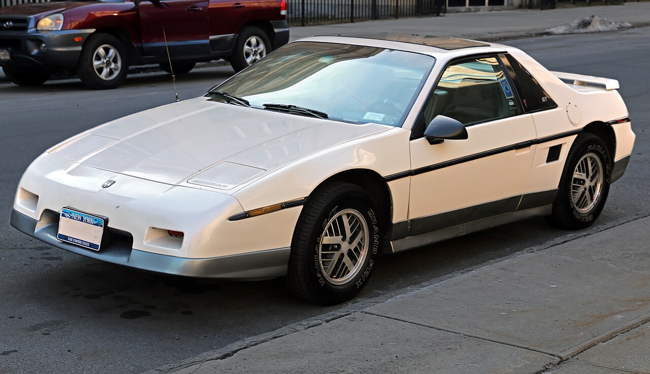 White 1985 Pontiac Fiero GT with a rear spoiler, front left three-quarter view parked beside a kerb.