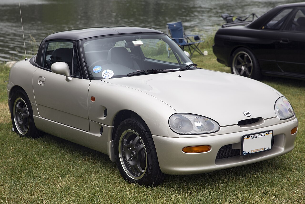 Pale silver 1991 Suzuki Cappuccino with a black hard top, front right three-quarter view on grass beside a lake.