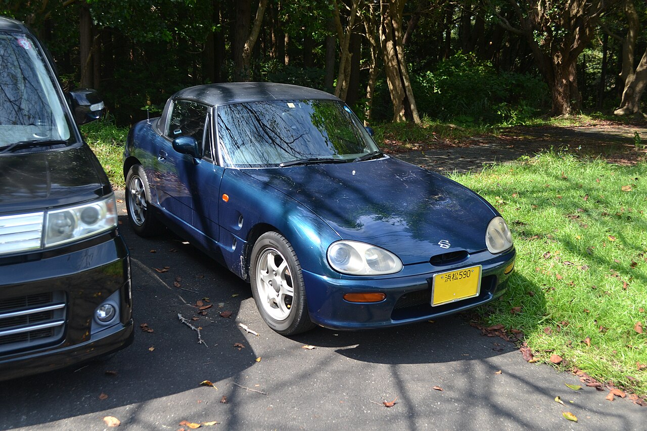 Dark blue Suzuki Cappuccino with a black hard top, front right three-quarter view parked on tarmac beside trees.