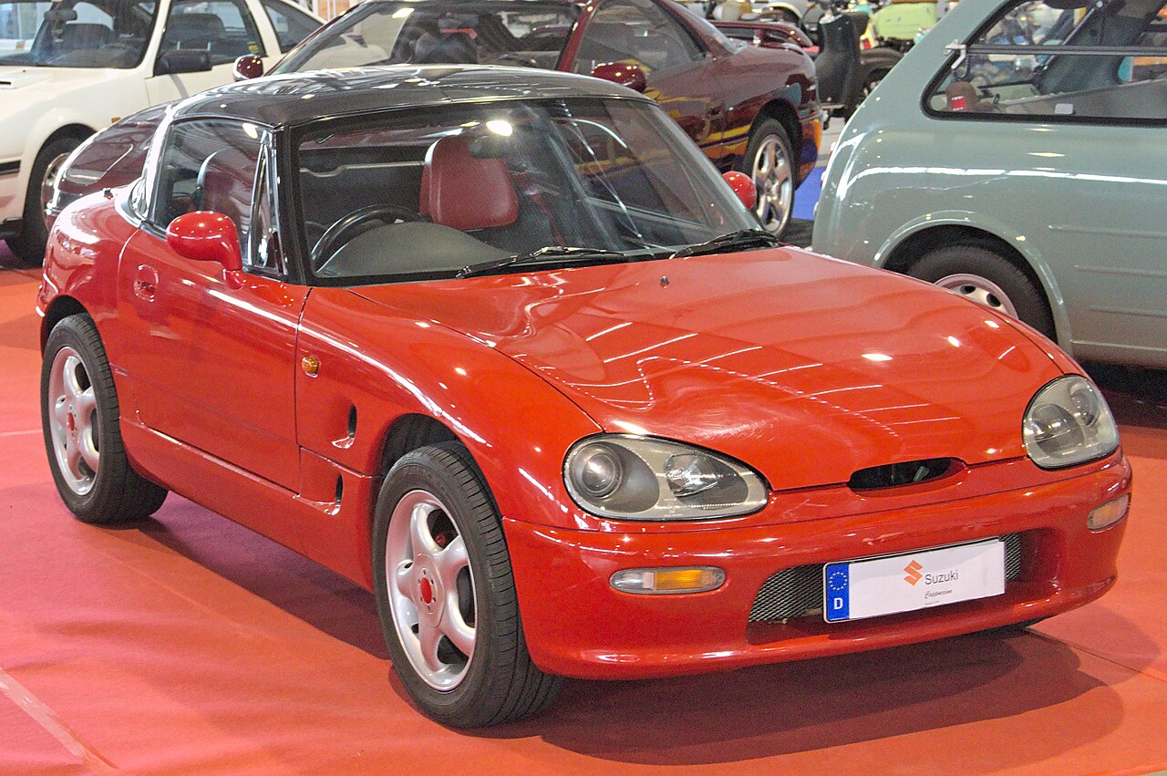 Red Suzuki Cappuccino with its black roof fitted, front three-quarter view on an indoor show floor.