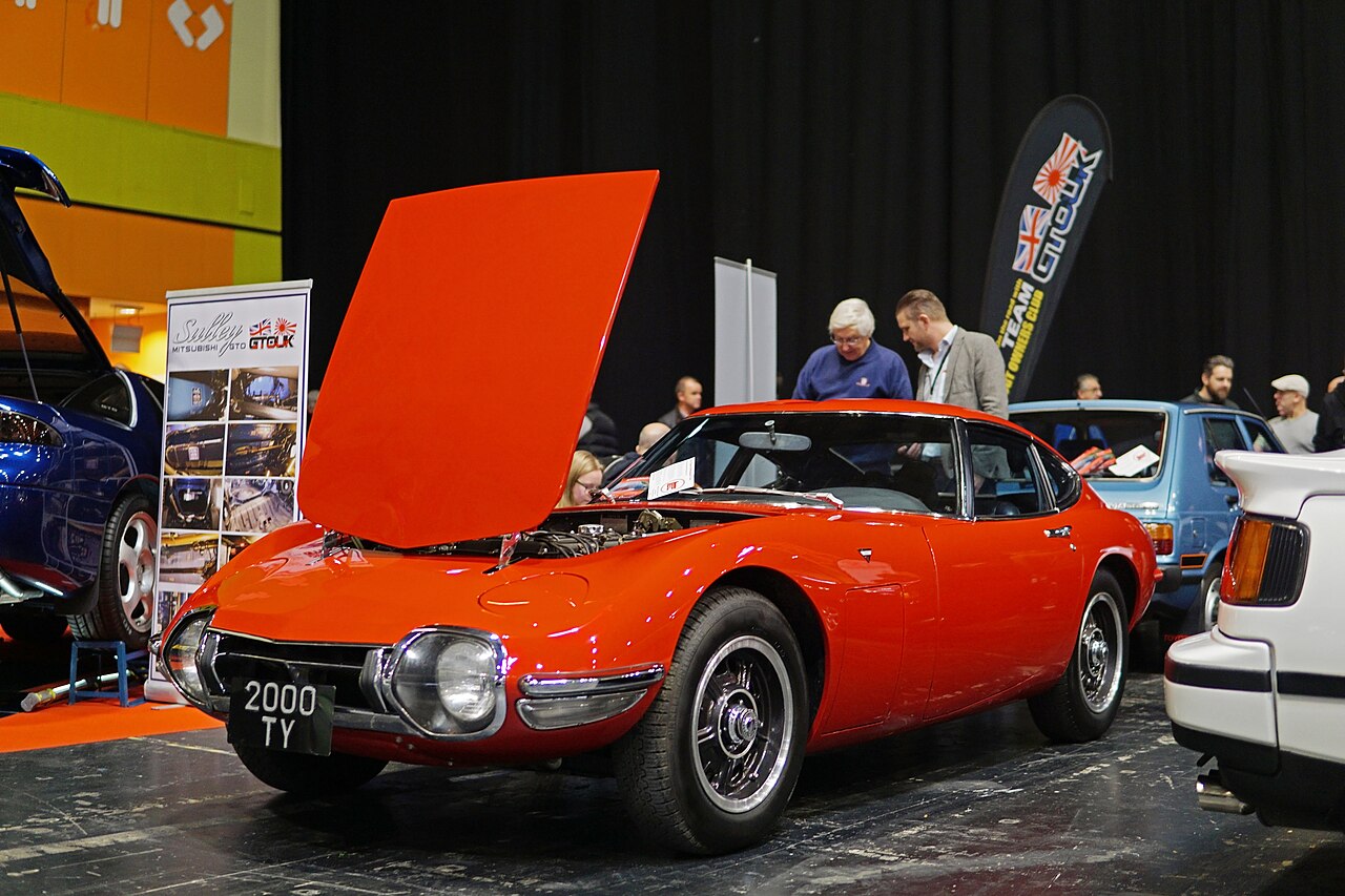 Red Toyota 2000GT coupe with its bonnet raised, front three-quarter view on a stand at an indoor classic car show.