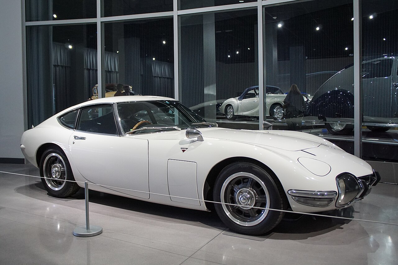 White Toyota 2000GT coupe, front right three-quarter view on a museum floor with a rope barrier in front.