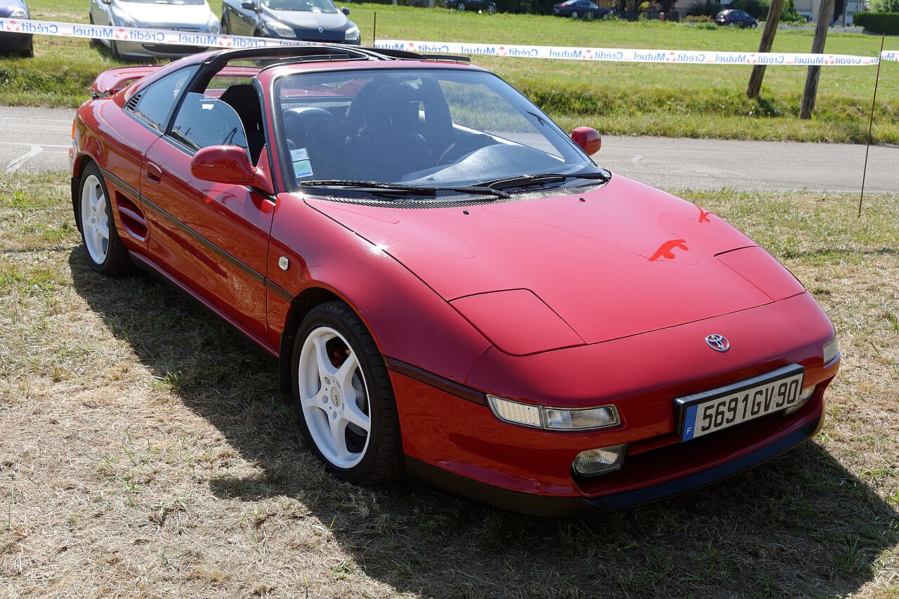 Red Toyota MR2 SW20 with its roof panels removed, front right three-quarter view on grass at a convertible show.
