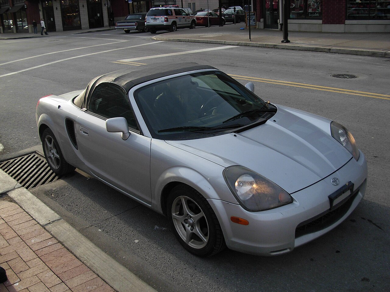 Silver Toyota MR2 Spyder with its black soft top up, front right three-quarter view from a raised angle on a city street.