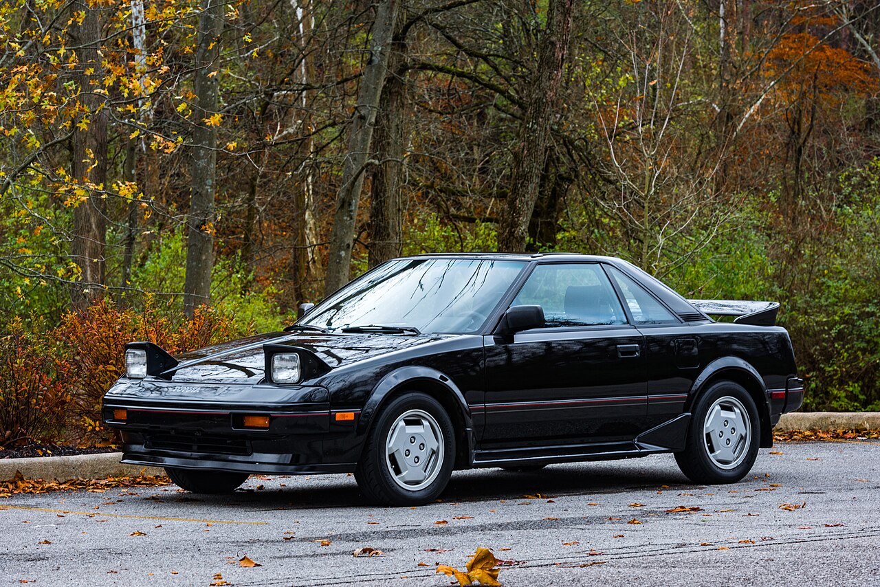 Black 1986 first-generation Toyota MR2 with its pop-up headlights raised, front three-quarter view in an autumn car park.