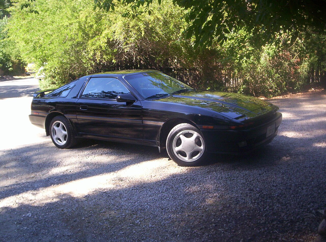 Black 1988 Toyota Supra Turbo, front three-quarter view parked on a gravel drive under trees.