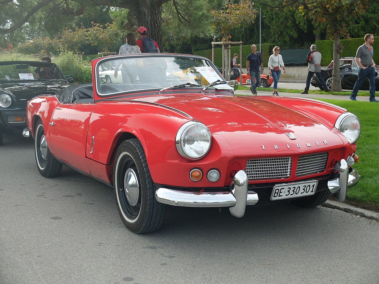Red Triumph Spitfire 4 with its roof down and white-wall tyres, front three-quarter view on a street at a classic car meeting.