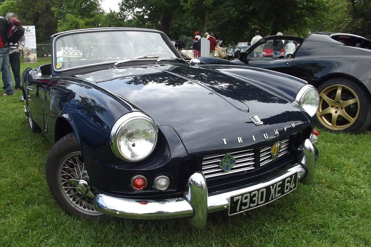 Dark blue Triumph Spitfire Mk2 with wire wheels and its roof down, front three-quarter view on grass at an outdoor car show.