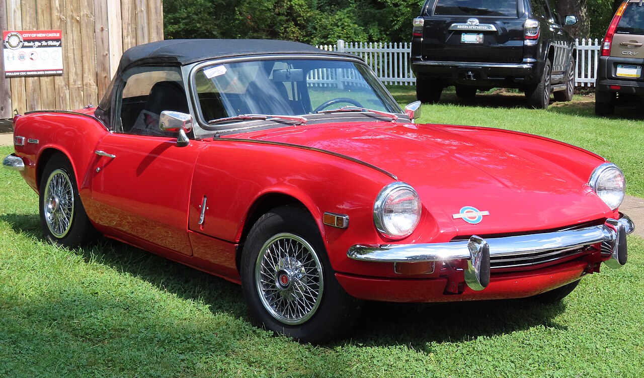 Red 1970 Triumph Spitfire Mk3 with its black soft top up and wire wheels, front three-quarter view on grass at a car show.