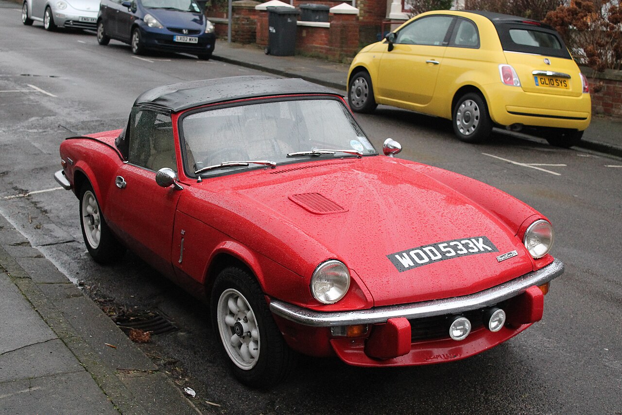 Red 1972 Triumph Spitfire Mk4 with its black soft top up and white alloy wheels, front three-quarter view on a wet street.