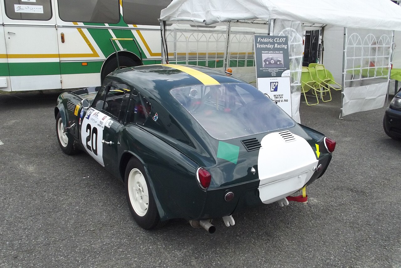 1965 Triumph Spitfire Le Mans racing coupe in dark green with a yellow roof stripe, rear three-quarter view in a paddock.