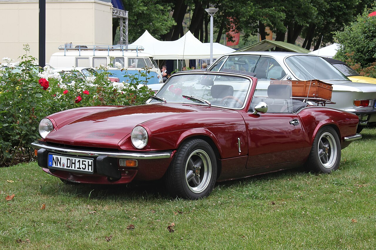Maroon Triumph Spitfire 1500 with its roof down and a brown suitcase on a boot rack, front three-quarter view on grass.