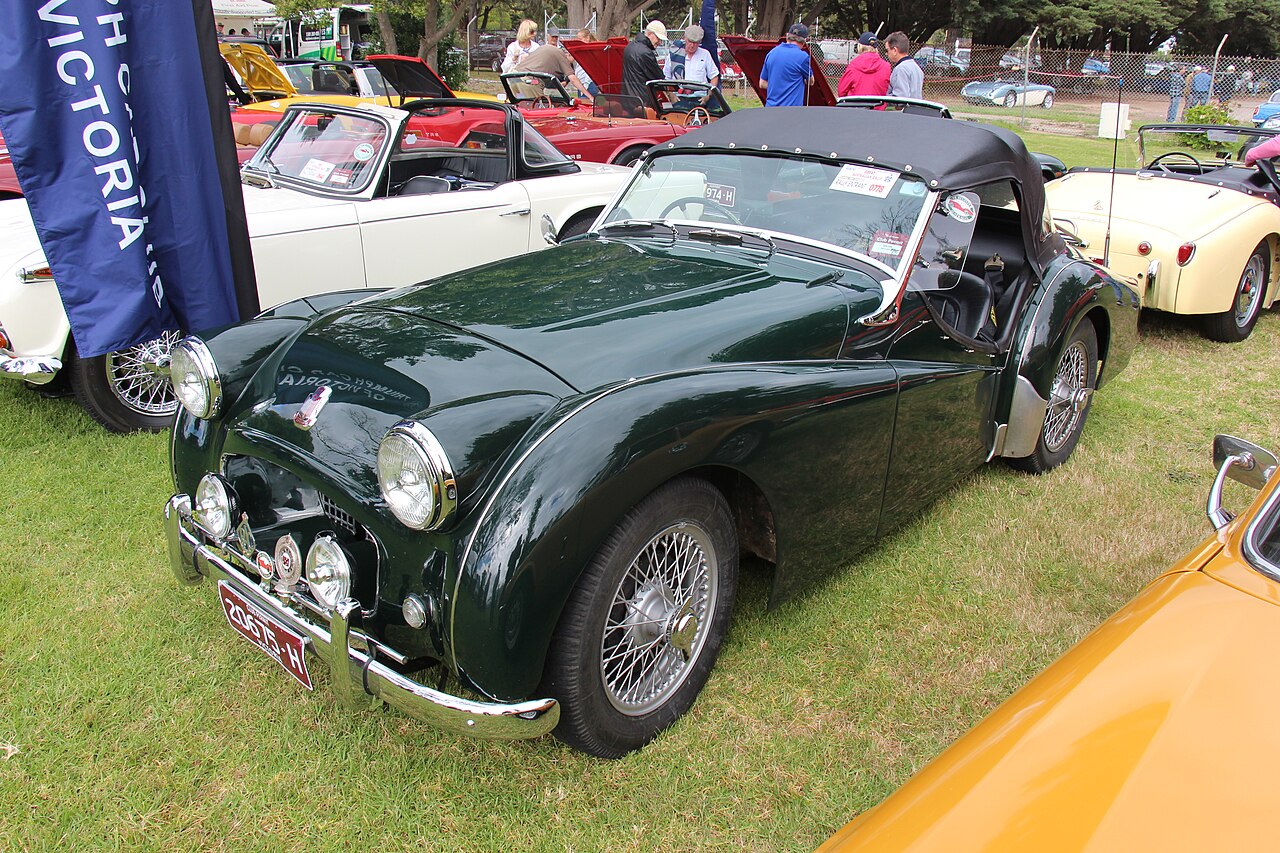 Dark green 1954 Triumph TR2 with its black soft top up and wire wheels, front left three-quarter view on grass at a car show.
