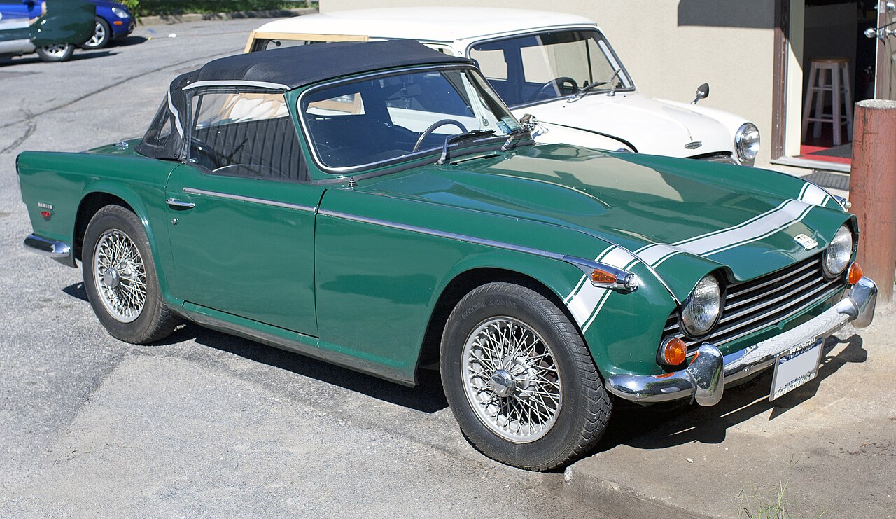Green 1967 Triumph TR250 with white bonnet stripes and its soft top up, front right three-quarter view in a car park.