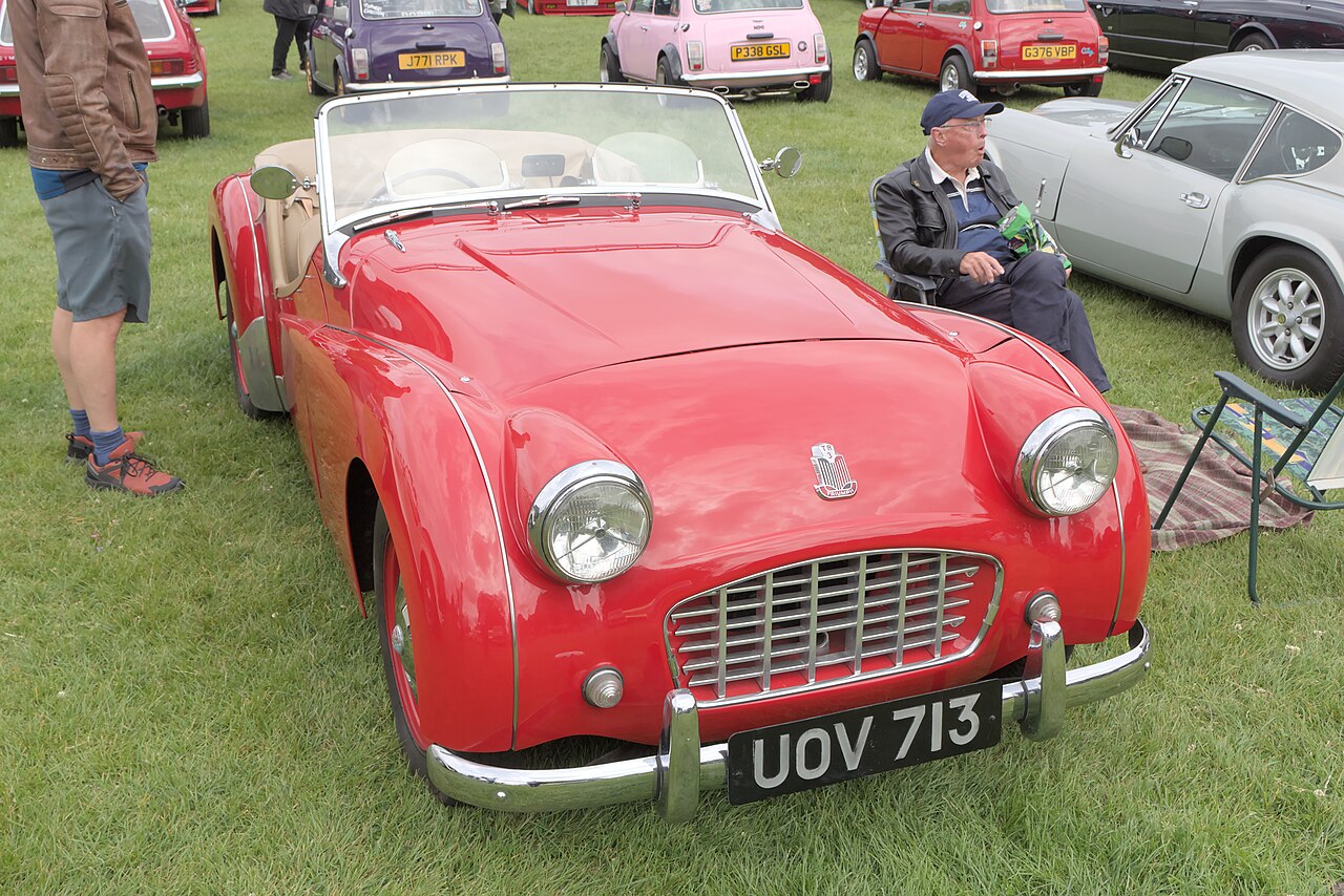 Red Triumph TR3 with its roof down and a tan interior, front three-quarter view on grass at a classic car show.