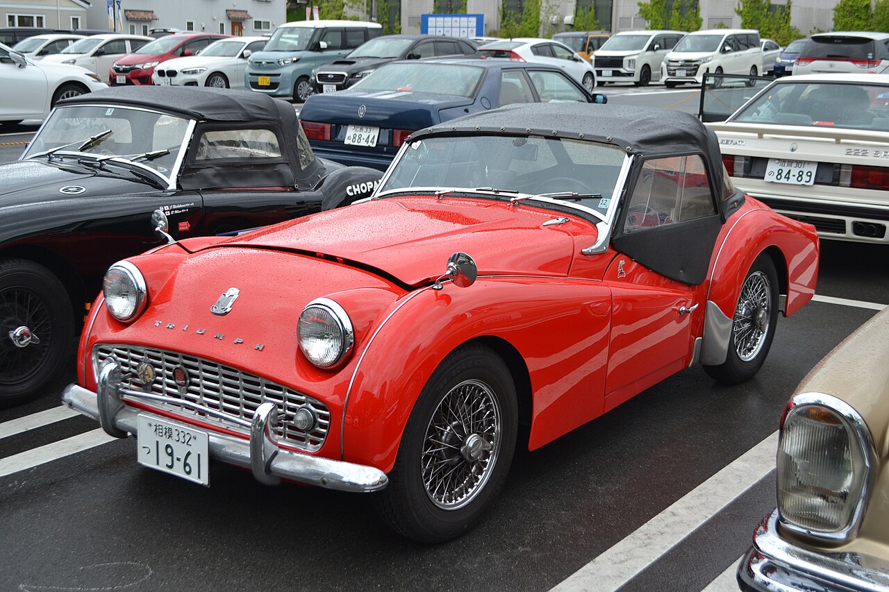 Red Triumph TR3A roadster with its black soft top fitted, front three-quarter view in a car park.