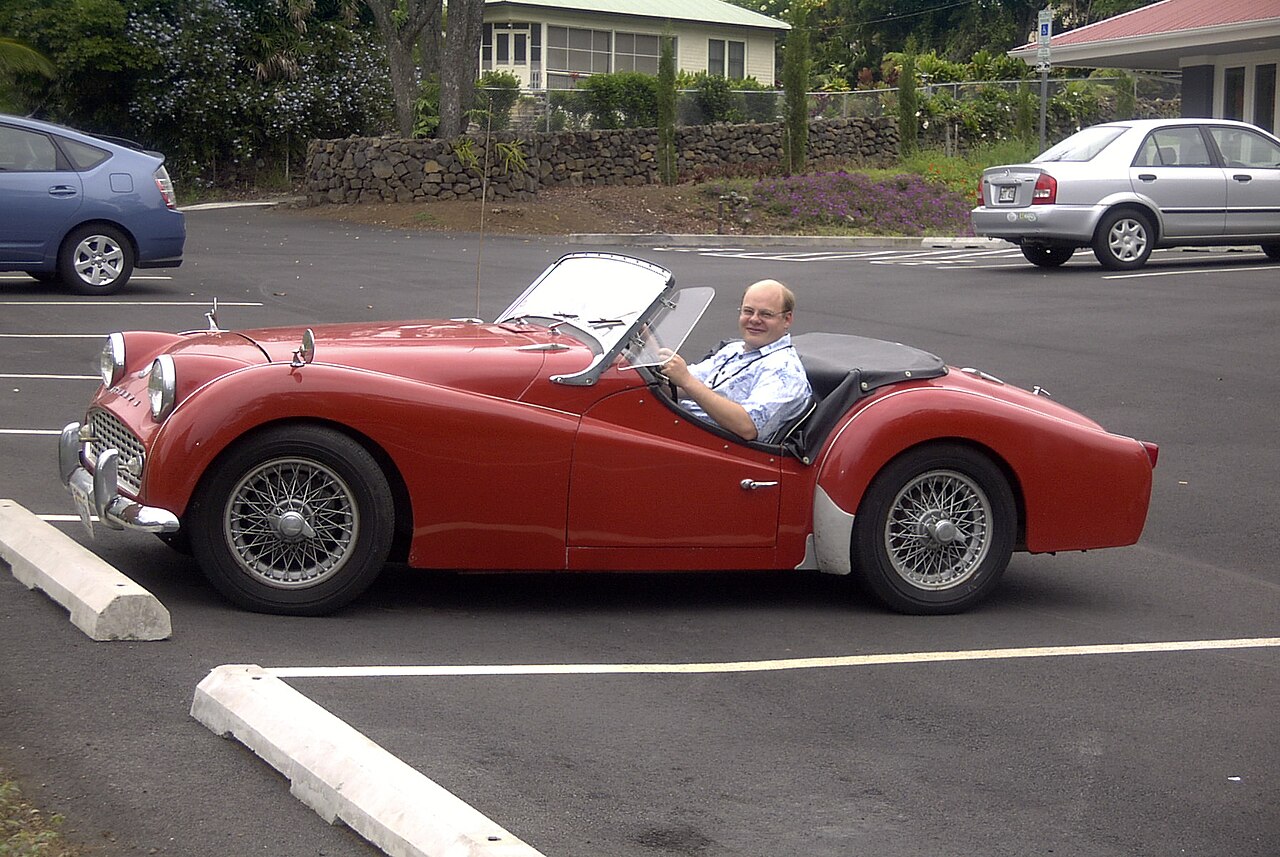 Red 1962 Triumph TR3B with wire wheels, roof down and a man at the wheel, side-on from the left in a car park.