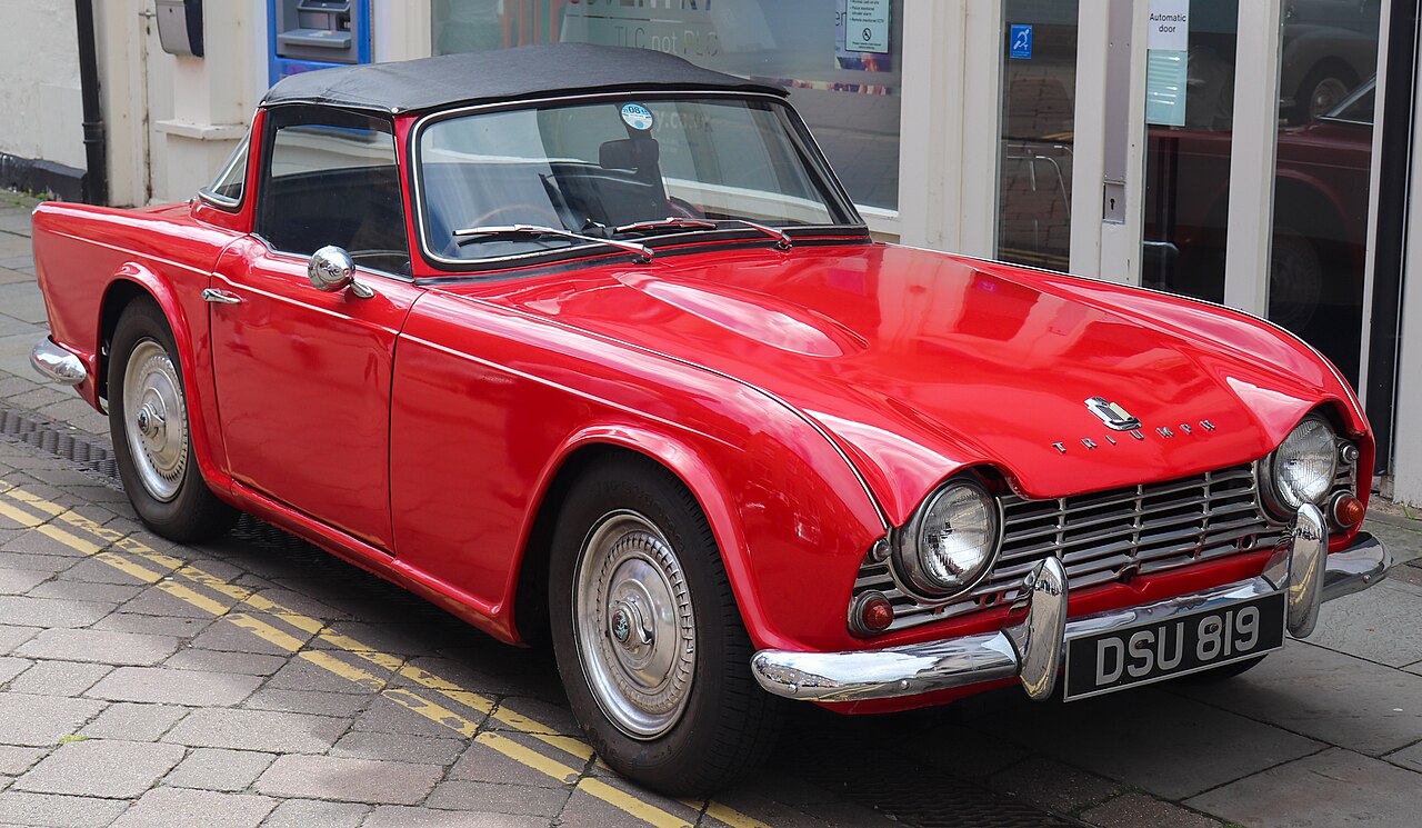Red 1962 Triumph TR4 with a black roof, front three-quarter view parked on a paved street beside yellow lines.