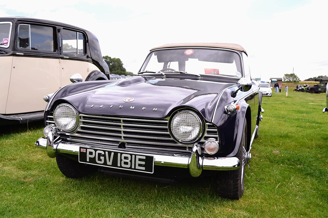 Dark blue 1967 Triumph TR4A with its tan roof up, shown almost head-on on grass beside a vintage saloon at a car show.