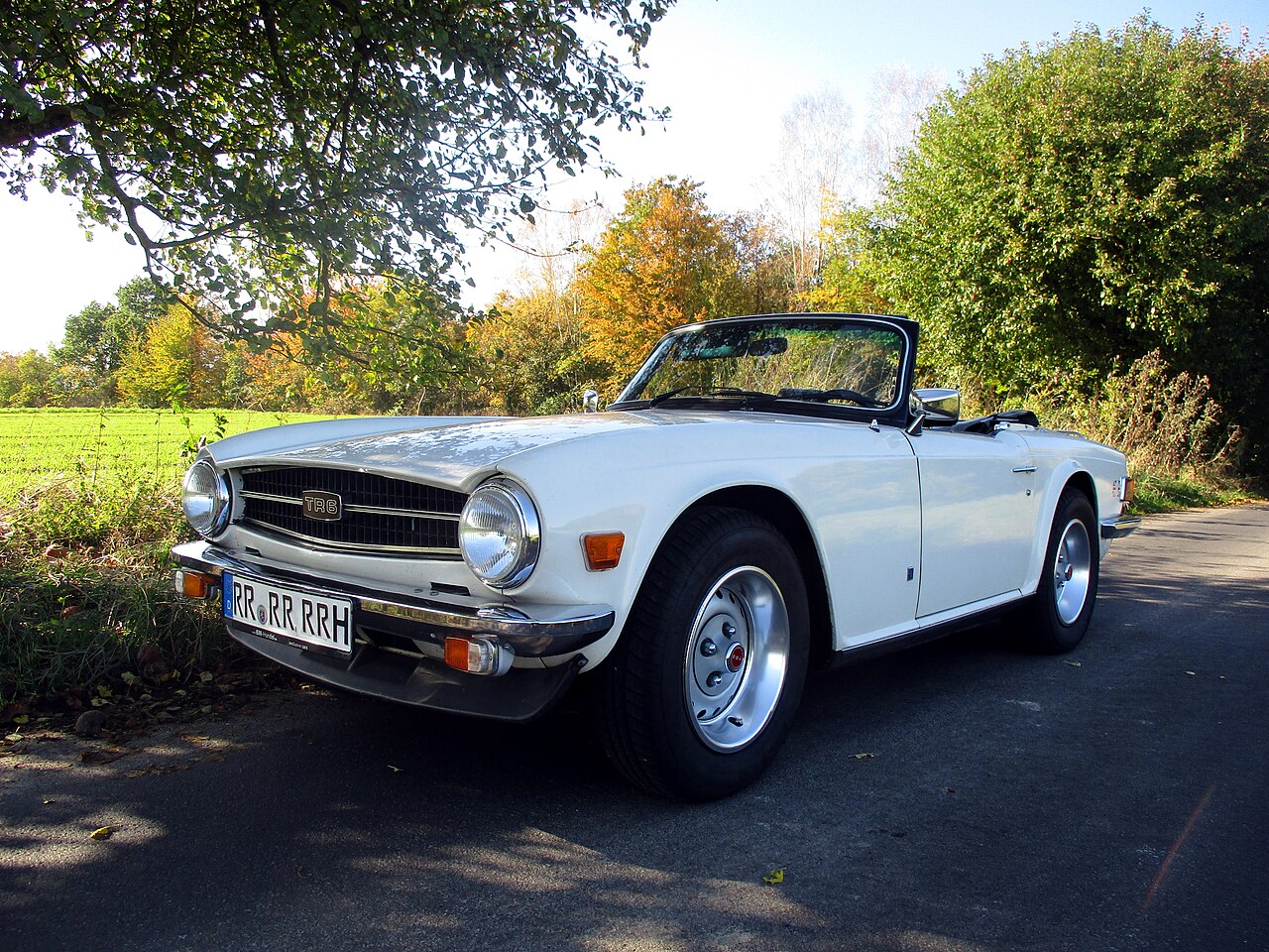 White Triumph TR6 roadster with its roof down, front three-quarter view on a country lane lined with autumn trees.