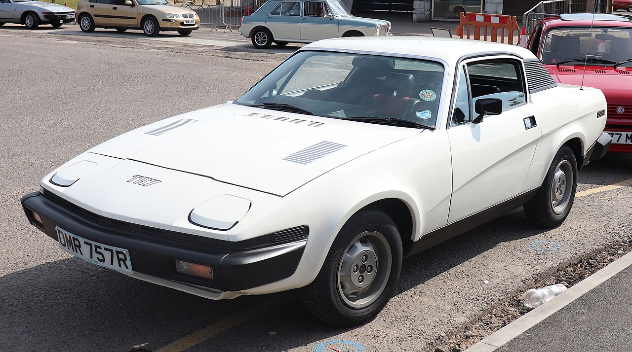White 1977 Triumph TR7 coupe with silver steel wheels, front three-quarter view in a car park with other cars behind.