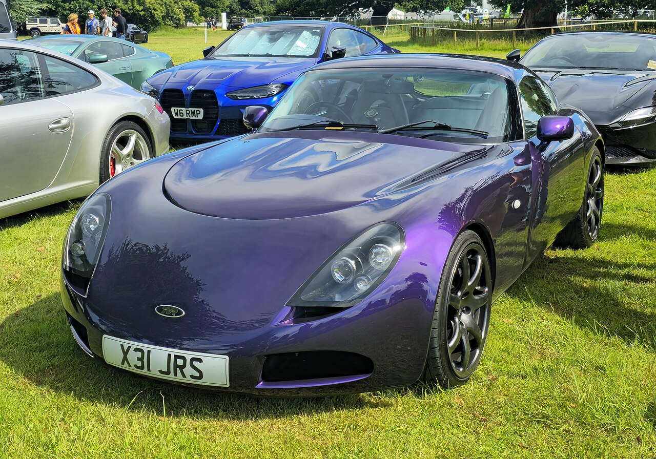 Purple TVR T350C coupe on grass at a car show, front left three-quarter view with other cars parked behind.